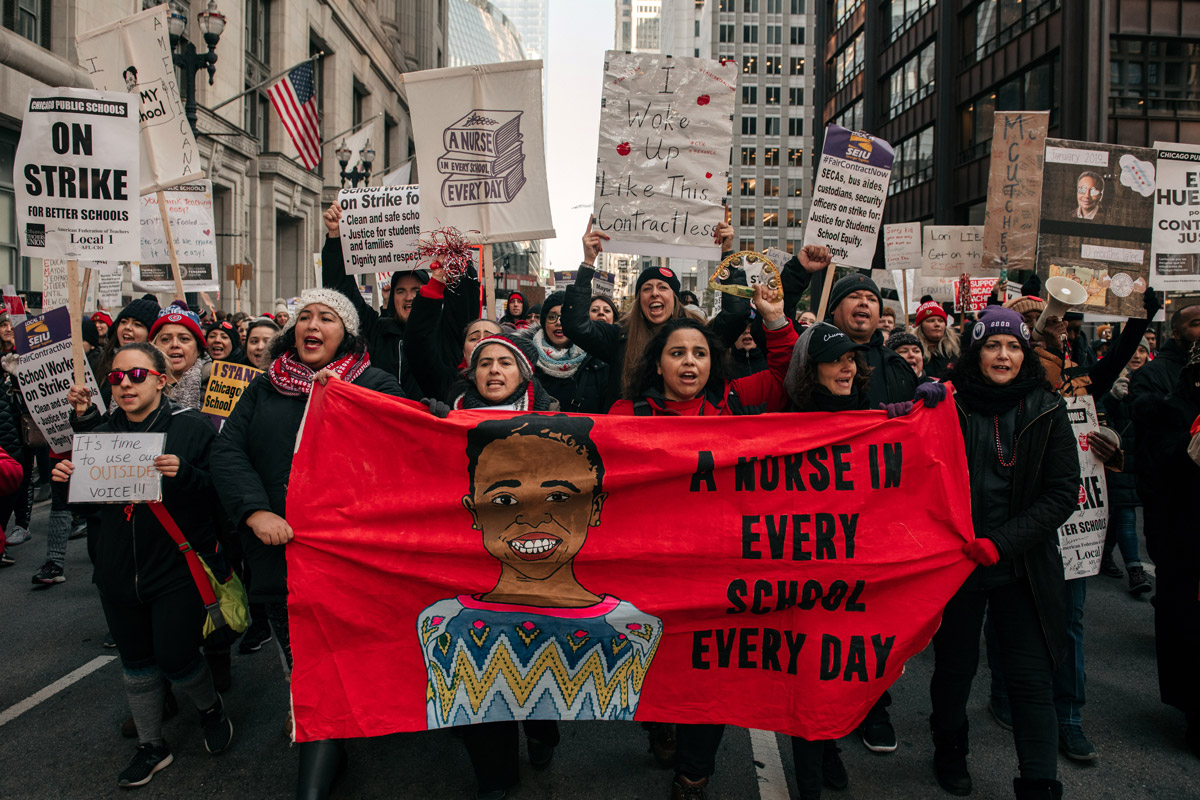 Striking Chicago teachers marching in the street, carrying a banner "A Nurse in Every School, Every Day."
